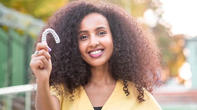 A smiling woman with curly hair, holding a white object in her hand against a blurred background.