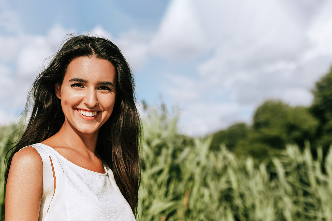 A young woman with long hair is standing in a field of tall crops, smiling towards the camera.