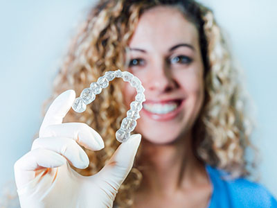 A woman wearing a blue surgical mask holds up a large, clear plastic dental retainer.