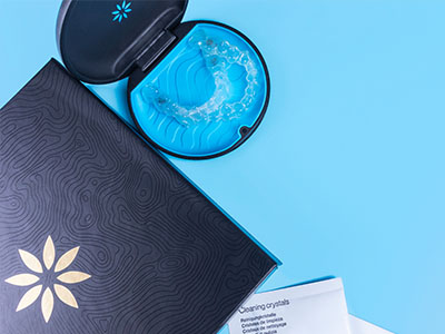 The image shows a blue toothbrush with bristles, placed inside an open, black case that resembles a jewelry box. In the background, there is a closed book or notebook with a floral design on its cover and a small, decorative object resembling a starfish or flower in front of it. The overall style of the image suggests a clean, minimalist aesthetic with a focus on personal care products.