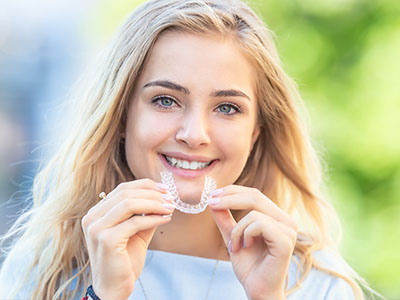 A young woman with a radiant smile is holding up a clear, plastic retainer. She appears to be outdoors, possibly in a park or garden setting.