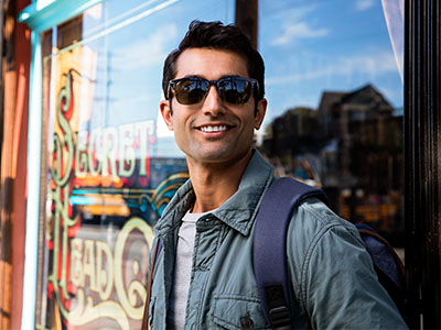 The image features a man standing in front of a storefront window, wearing sunglasses and a backpack, with a smile on his face.